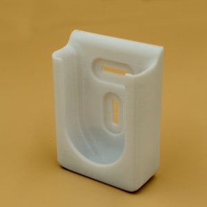 May include: A white, 3D-printed plastic wall mount with a rectangular base and a curved top. It features two oval cutouts and a rectangular opening. The mount is designed to hold an item, likely a phone or small device.