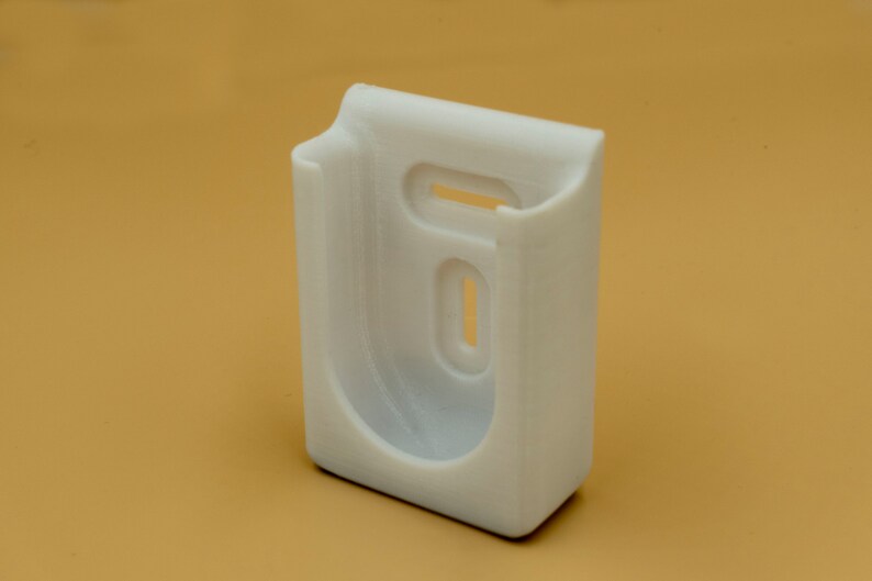 May include: A white, 3D-printed plastic wall mount with a rectangular base and a curved top. It features two oval cutouts and a rectangular opening. The mount is designed to hold an item, likely a phone or small device.