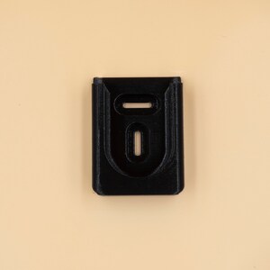 May include: A black, rectangular object with two oval cutouts and a horizontal slot at the top. The object is centered on a light tan background. The item appears to be a holder or mount.