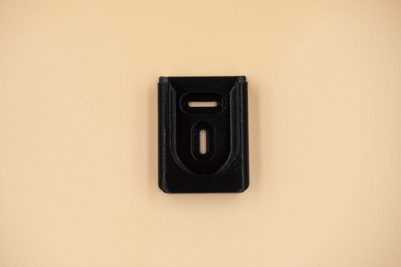 May include: A black, rectangular object with two oval cutouts and a horizontal slot at the top. The object is centered on a light tan background. The item appears to be a holder or mount.