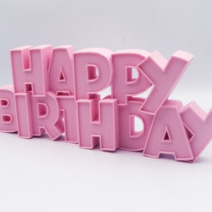 May include: A pink, three-dimensional "HAPPY BIRTHDAY" sign. The letters are in a bold, sans-serif font and are arranged in two stacked lines. The sign is made of a solid material and is set against a white background.