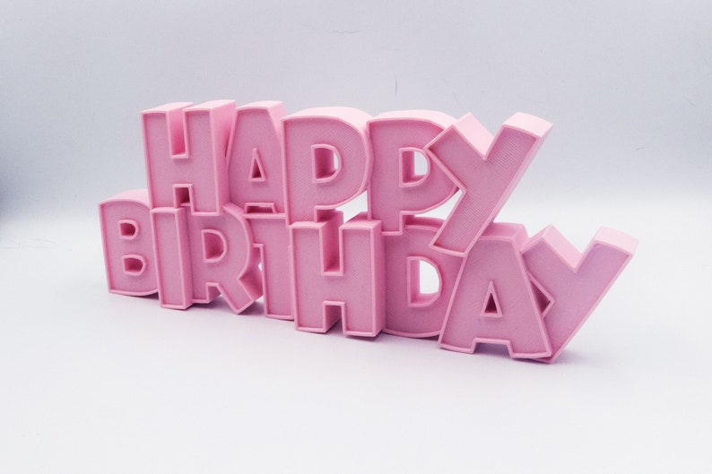 May include: A pink, three-dimensional "HAPPY BIRTHDAY" sign. The letters are in a bold, sans-serif font and are arranged in two stacked lines. The sign is made of a solid material and is set against a white background.