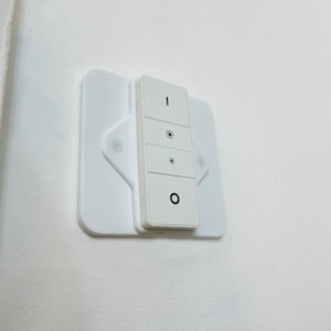 Puede incluir: Un interruptor de luz inteligente blanco con cuatro botones y un diseño rectangular. El interruptor está montado en una pared blanca y tiene símbolos para encendido/apagado y control de brillo.