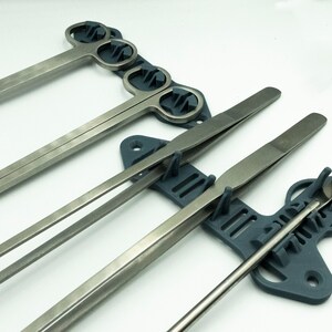 Pode incluir: Um suporte de ferramentas de pl&aacute;stico cinza com cinco ranhuras para segurar v&aacute;rias ferramentas. O suporte tem um design &uacute;nico com uma forma curva e uma superf&iacute;cie texturizada. As ferramentas s&atilde;o de metal e incluem um par de pin&ccedil;as, um par de f&oacute;rceps e tr&ecirc;s esp&aacute;tulas.