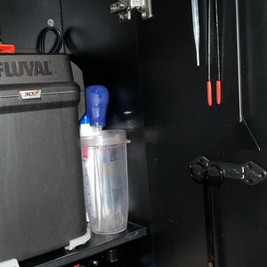 Pode incluir: Um arm&aacute;rio preto com um filtro Fluval 307, um recipiente de pl&aacute;stico transparente com tampa azul e uma garrafa de inibidor de algas de carbono f&aacute;cil de aqu&aacute;rio co-op. Tamb&eacute;m h&aacute; v&aacute;rias ferramentas penduradas no lado do arm&aacute;rio.