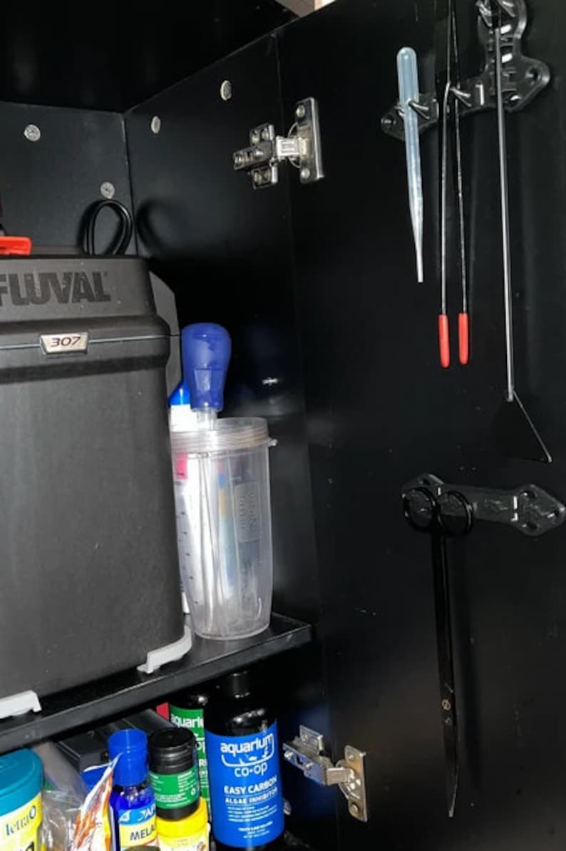 Pode incluir: Um arm&aacute;rio preto com um filtro Fluval 307, um recipiente de pl&aacute;stico transparente com tampa azul e uma garrafa de inibidor de algas de carbono f&aacute;cil de aqu&aacute;rio co-op. Tamb&eacute;m h&aacute; v&aacute;rias ferramentas penduradas no lado do arm&aacute;rio.