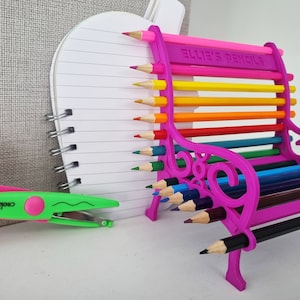 May include: A pink plastic pencil holder shaped like a bench with the words "Ellie's Pencils" on the back. The bench is filled with colored pencils.