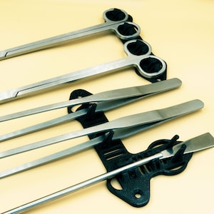 Pode incluir: Um conjunto de cinco pin&ccedil;as prateadas e uma pequena esp&aacute;tula prateada s&atilde;o exibidas em um suporte de pl&aacute;stico preto. O suporte tem um design &uacute;nico com v&aacute;rias ranhuras para segurar as ferramentas.