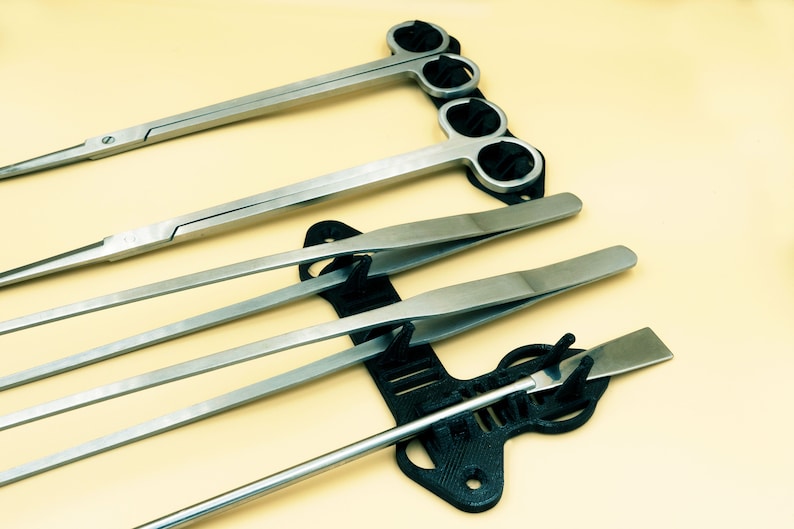 Pode incluir: Um conjunto de cinco pin&ccedil;as prateadas e uma pequena esp&aacute;tula prateada s&atilde;o exibidas em um suporte de pl&aacute;stico preto. O suporte tem um design &uacute;nico com v&aacute;rias ranhuras para segurar as ferramentas.