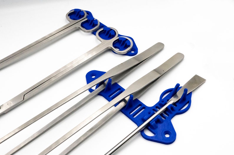 Pode incluir: Um conjunto de cinco ferramentas cir&uacute;rgicas com al&ccedil;as de pl&aacute;stico azul, incluindo pin&ccedil;as, f&oacute;rceps e uma esp&aacute;tula, s&atilde;o exibidas em um suporte de pl&aacute;stico azul.