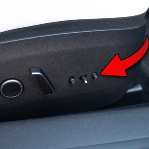 Op de afbeelding: Close-up van een zwarte autostoel met een zilveren bedieningspaneel. Het paneel heeft een ronde knop, een hendel en vier kleine knoppen. De knoppen zijn gelabeld met symbolen voor de stoelverstelling.