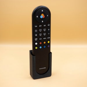 May include: A black remote control with multiple buttons and a color wheel, resting in a matching black holder. The remote has the brand name "novastella" printed on the holder. The background is a solid light orange.