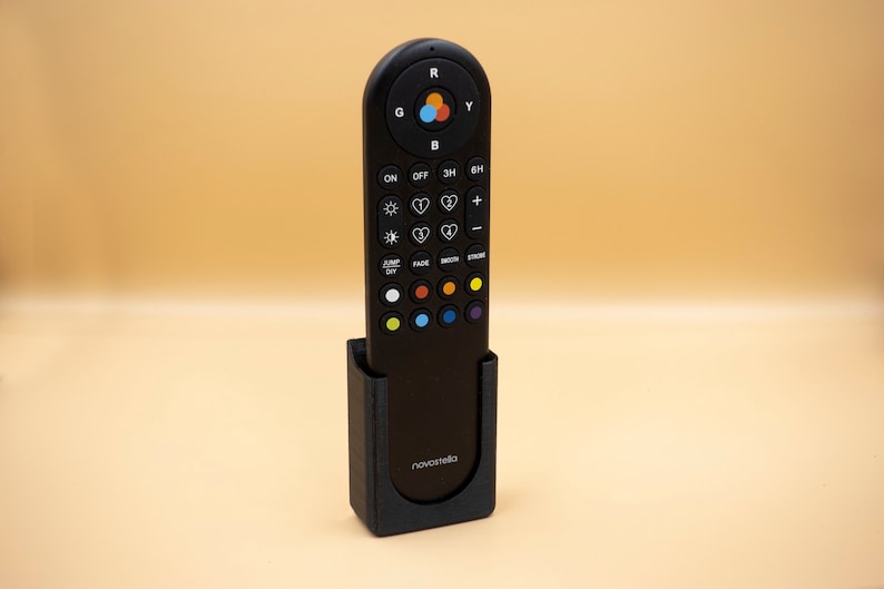 May include: A black remote control with multiple buttons and a color wheel, resting in a matching black holder. The remote has the brand name "novastella" printed on the holder. The background is a solid light orange.