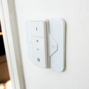 Può includere: Un interruttore di luce intelligente bianco montato su una parete bianca. L'interruttore ha quattro pulsanti con simboli per l'alimentazione, la luminosità e l'accensione/spegnimento. L'interruttore è rettangolare con angoli arrotondati.