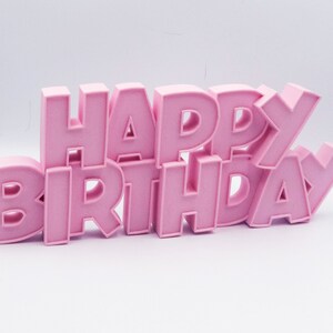 May include: A pink "HAPPY BIRTHDAY" sign. The letters are in a bold, blocky font and are arranged in a slightly staggered, three-dimensional design. The sign is a solid, light pink color and sits on a white surface.