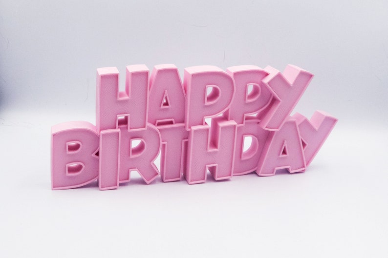 May include: A pink "HAPPY BIRTHDAY" sign. The letters are in a bold, blocky font and are arranged in a slightly staggered, three-dimensional design. The sign is a solid, light pink color and sits on a white surface.