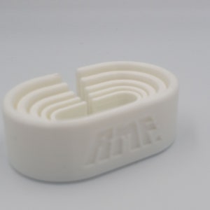 May include: A white, oval-shaped object with a series of nested, curved slots. The object has a smooth surface and a cut-out section. The letters "AME" are embossed on the side. The object appears to be a tool or a holder.
