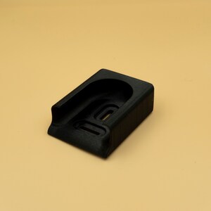 May include: A black, rectangular object with a curved top and a recessed area. The object has a textured surface and appears to be made of a solid material. The background is a solid tan color.