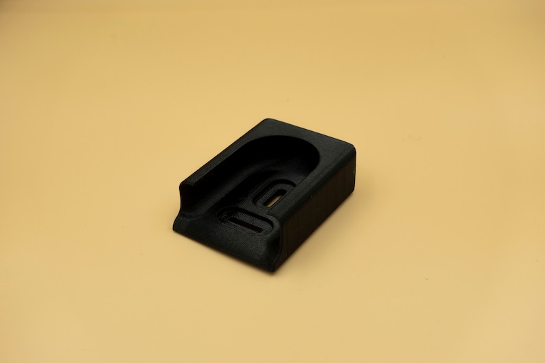 May include: A black, rectangular object with a curved top and a recessed area. The object has a textured surface and appears to be made of a solid material. The background is a solid tan color.