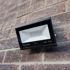 May include: A black rectangular LED floodlight mounted on a red brick wall. The floodlight has a white interior with a grid of small LED lights. The text "novostella" is visible on the light. The text "25W IP66" is also visible.