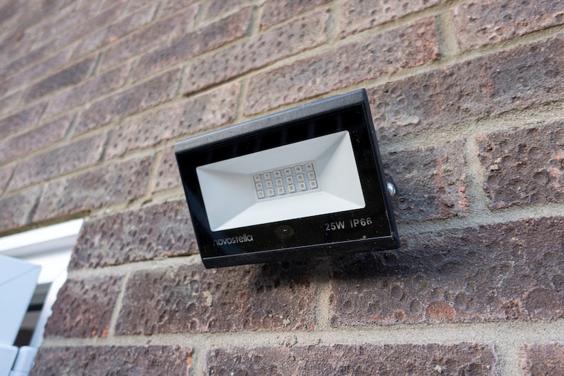 May include: A black rectangular LED floodlight mounted on a red brick wall. The floodlight has a white interior with a grid of small LED lights. The text "novostella" is visible on the light. The text "25W IP66" is also visible.