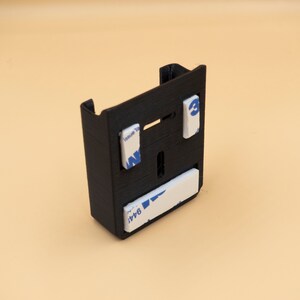 May include: A black, rectangular 3D-printed bracket with a slot and two tabs at the top. It has three white adhesive pads with blue text. The bracket is designed to hold an item.