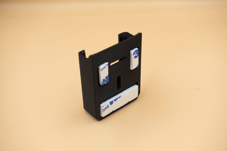May include: A black, rectangular 3D-printed bracket with a slot and two tabs at the top. It has three white adhesive pads with blue text. The bracket is designed to hold an item.