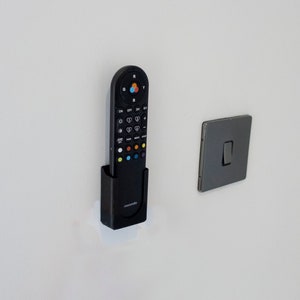May include: A black remote control in a black holder fixed to a white wall, beside a grey light switch. The remote features various buttons with coloured accents and the brand name "novosonic".