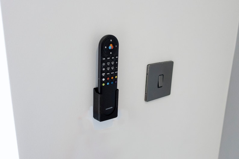 May include: A black remote control in a black holder mounted on a white wall, next to a gray light switch. The remote has multiple buttons with colorful accents and the brand name "novosonic".