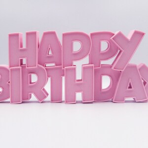 May include: Pink, three-dimensional letters spell out "HAPPY BIRTHDAY" against a white background. The letters are arranged in a stacked, slightly angled design, creating a celebratory visual.