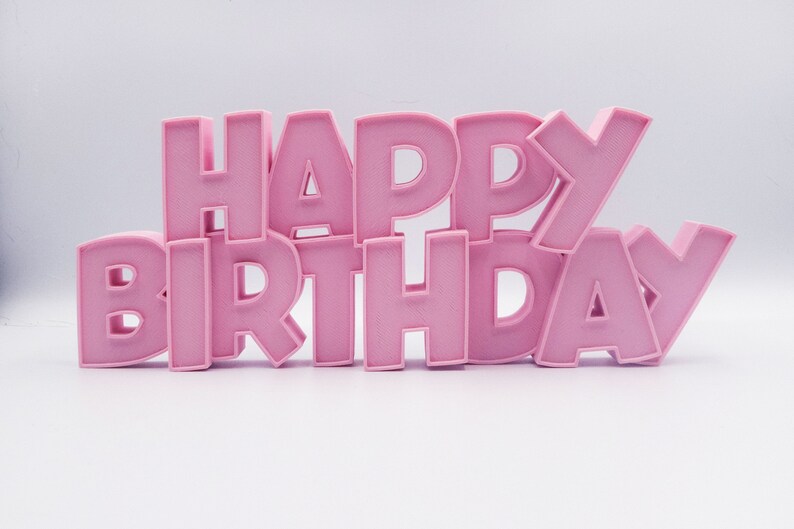 May include: Pink, three-dimensional letters spell out "HAPPY BIRTHDAY" against a white background. The letters are arranged in a stacked, slightly angled design, creating a celebratory visual.