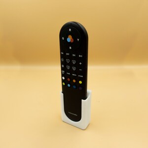 May include: A black remote control with multiple buttons and a color wheel, resting in a white holder. The remote has buttons labeled with symbols and text. The holder has the word "Astromedia" printed on it.
