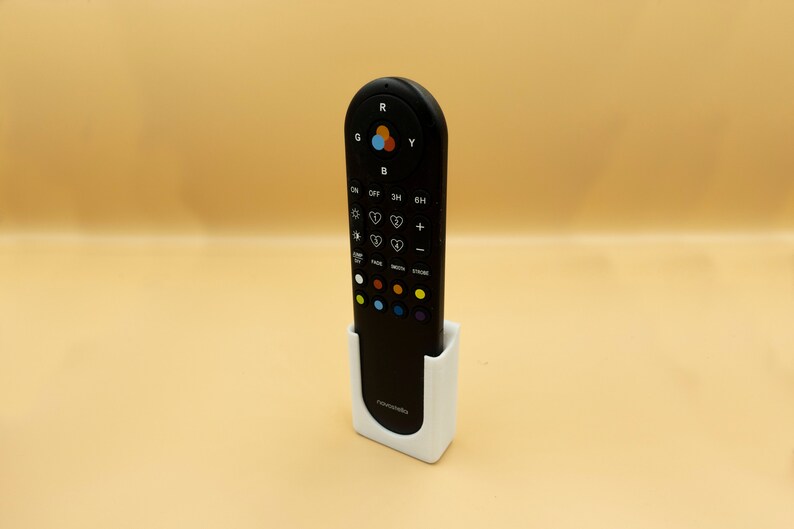 May include: A black remote control with multiple buttons and a color wheel, resting in a white holder. The remote has buttons labeled with symbols and text. The holder has the word "Astromedia" printed on it.