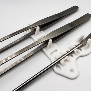 May include: A set of stainless steel laboratory tools, including tweezers, a spatula, and a stirring rod, resting in a white 3D-printed holder. The tools are arranged against a white background.