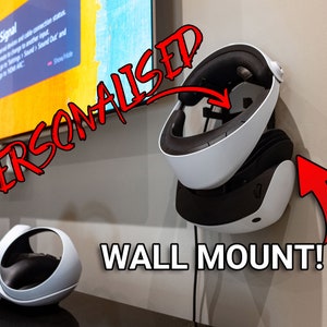 May include: A white and black virtual reality headset is mounted on a wall with a red arrow pointing to it. The text "PERSONALISED WALL MOUNT!" is written in red on the wall. A white and black virtual reality headset is sitting on a black surface.