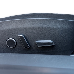 May include: Close-up of a black car seat with white upholstery. The seat features a round button and two silver-trimmed levers for adjusting the seat position. The seat is made of textured black material.