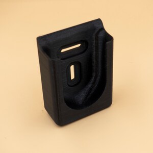 May include: A black, rectangular holder with a curved interior and two rectangular cutouts. The holder is made of a textured material and is designed to hold an item. The item is placed on a light tan background.