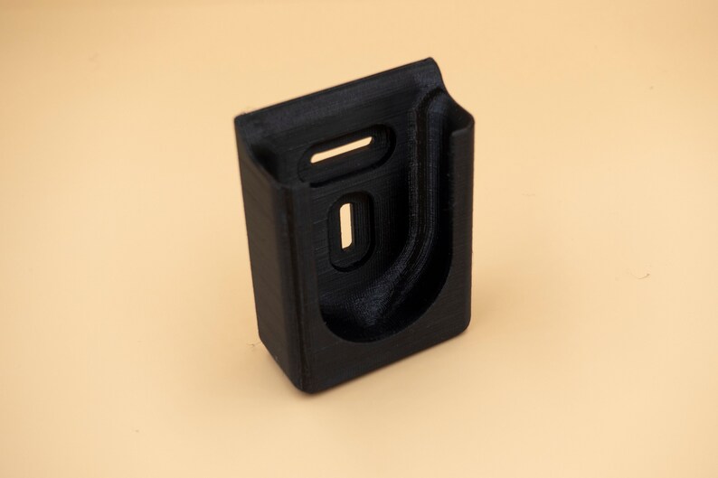 May include: A black, rectangular holder with a curved interior and two rectangular cutouts. The holder is made of a textured material and is designed to hold an item. The item is placed on a light tan background.