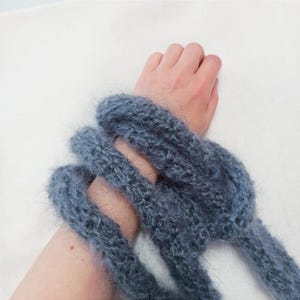 May include: A close-up of a blue knitted scarf wrapped around a wrist. The scarf is made of a soft, fuzzy yarn. The background is a plain white surface.