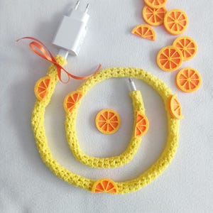 Puede incluir: Un envoltorio de cable de cargador de teléfono de crochet amarillo, decorado con rodajas de limón de fieltro naranja.