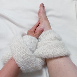 May include: Close-up of white, knitted leg warmers. The leg warmers are made of a soft, fluffy yarn and are worn on the lower legs. The image shows the texture of the knit and the cozy appearance of the leg warmers.
