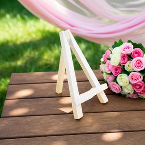 Könnte beinhalten: Ein kleines, weißes Holzstaffelei steht auf einer braunen Holzoberfläche. Rechts von der Staffelei befindet sich ein Strauß aus rosa und weißen Rosen. Im Hintergrund ist rosa Stoff drapiert, grünes Gras ist sichtbar.