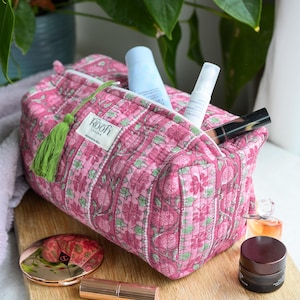 Może przedstawiać: Różowa, pikowana kosmetyczka w kwiaty z zielonym frędzlem, wypełniona produktami do pielęgnacji. Torba leży na drewnianej desce z pudrem, szminką i innymi kosmetykami do pielęgnacji skóry.