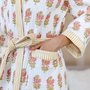 Könnte beinhalten: Weißer Steppmantel mit einem Blumenmuster in Rosa und Gelb. Der Bademantel hat eine gestreifte gelbe Bordüre und einen passenden Gürtel. Der Bademantel hat Taschen und ist in der Taille gebunden.
