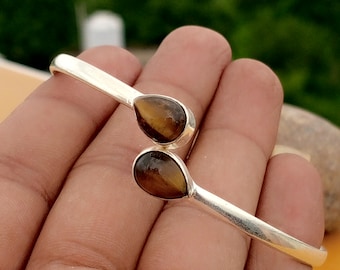May include: A silver bracelet featuring two teardrop-shaped tiger's eye stones. The bracelet has a simple, open design, with the stones set at each end. The tiger's eye stones are brown with golden streaks.