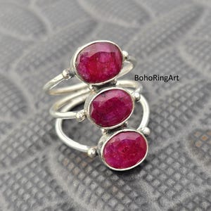 Może przedstawiać: Srebrny pierścionek z trzema owalnymi, głęboko czerwonymi kamieniami szlachetnymi. Pierścionek ma warstwowy design, z każdym kamieniem osadzonym na oddzielnej obrączce. Kamienie szlachetne są centralnym punktem, na tle teksturowanego szarego tła. Pierścionek to biżuteria.