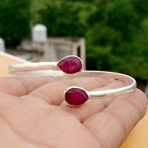 Könnte beinhalten: Ein silberfarbenes Armreif-Armband mit zwei tropfenförmigen, tiefroten Edelsteinen. Das Armband hat ein schlichtes, offenes Design, wobei die Edelsteine an jedem Ende eingesetzt sind. Das Armband wird auf einer Hand gezeigt.