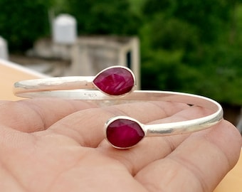 Puede incluir: Una pulsera abierta de plata con dos piedras preciosas de color rojo rosado intenso en forma de lágrima en cada extremo. La banda de metal es lisa y pulida, con un pequeño sello 925 grabado en la superficie interior, indicando plata de ley.