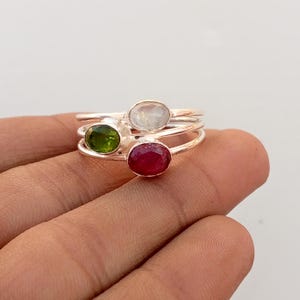 May include: A stackable ring set featuring three rings with oval gemstones. The gemstones are a vibrant green, a clear white, and a deep red. The rings are made of silver-colored metal and are displayed on a hand.