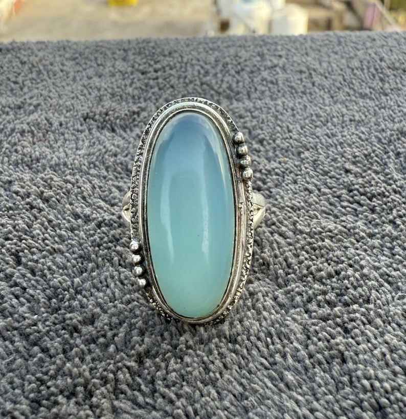 K&ouml;nnte beinhalten: Silberner Ring mit einem ovalen, hellblauen Edelstein. Der Stein ist in einem detaillierten Silberband mit dekorativen Elementen gefasst. Der Ring wird auf einer strukturierten, grauen Oberfl&auml;che pr&auml;sentiert. Der Edelstein ist ca. 2,5 cm lang.
